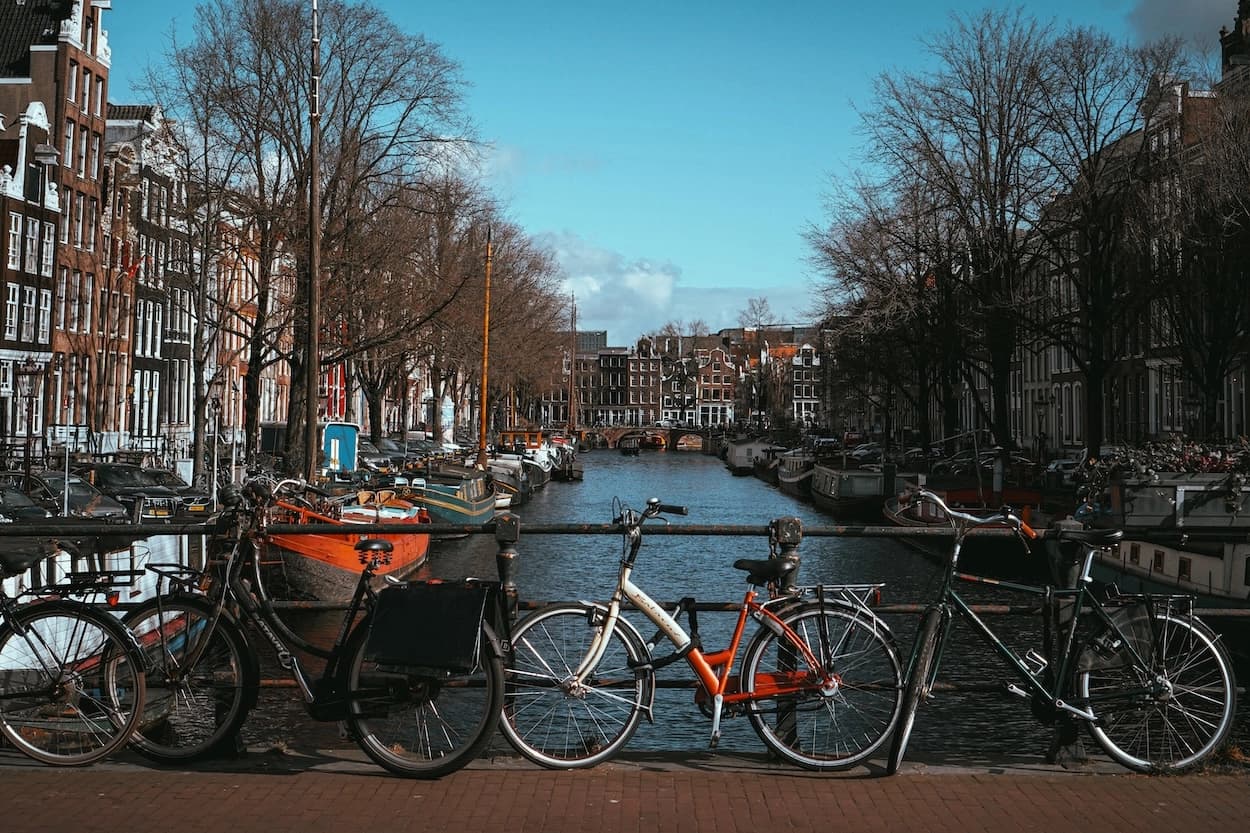 Bicycles chained to a bridge railing above an Amsterdam canal, houseboats and a barge moored along both banks under bare winter trees and a clear blue sky