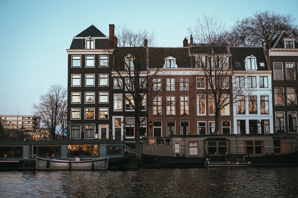 A row of tall canal houses at dusk seen across the water, lit windows reflecting the last of the light, with houseboats moored along the quay in front