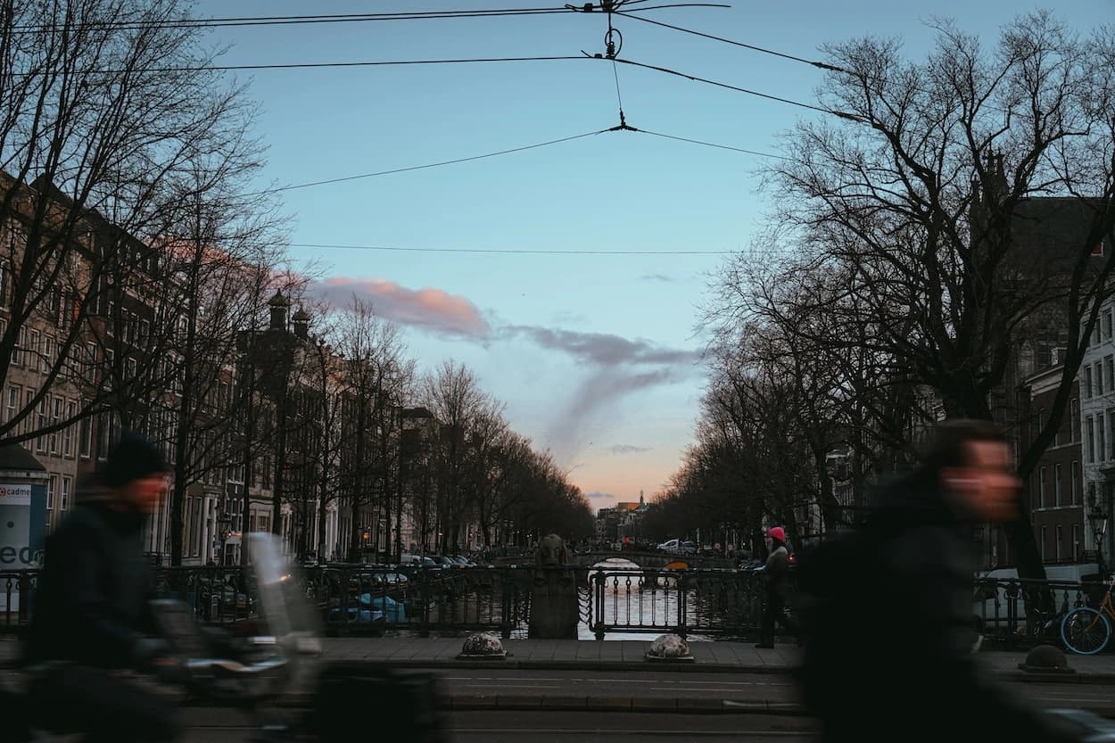 Two cyclists blurred past the camera on a canal bridge at dusk, the water and bare trees running away to a pink cloud over the rooftops behind them