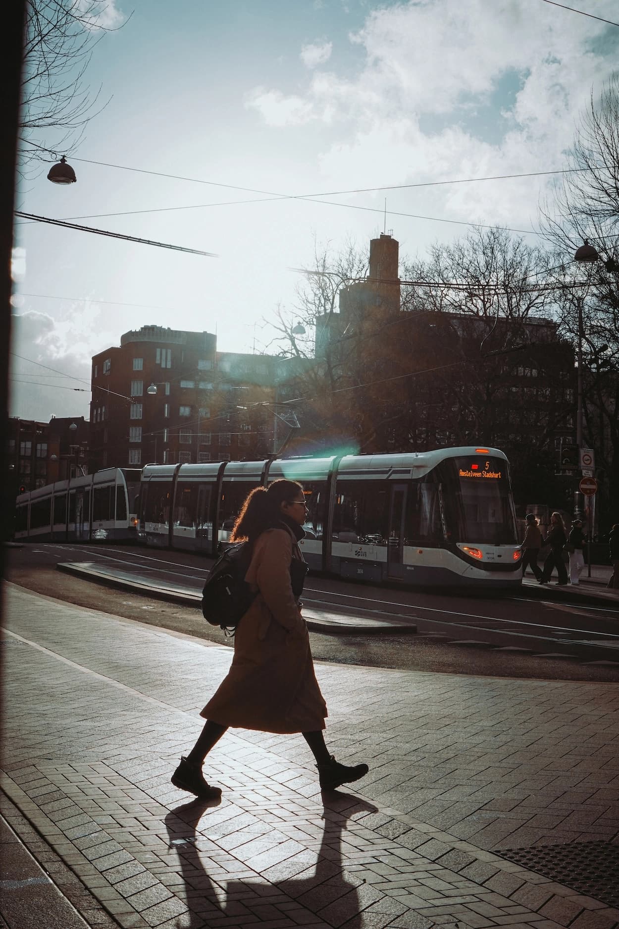 A woman in a long tan coat with a backpack walking a brick pavement into low winter sun, her shadow stretched behind her and a tram passing on the road beyond