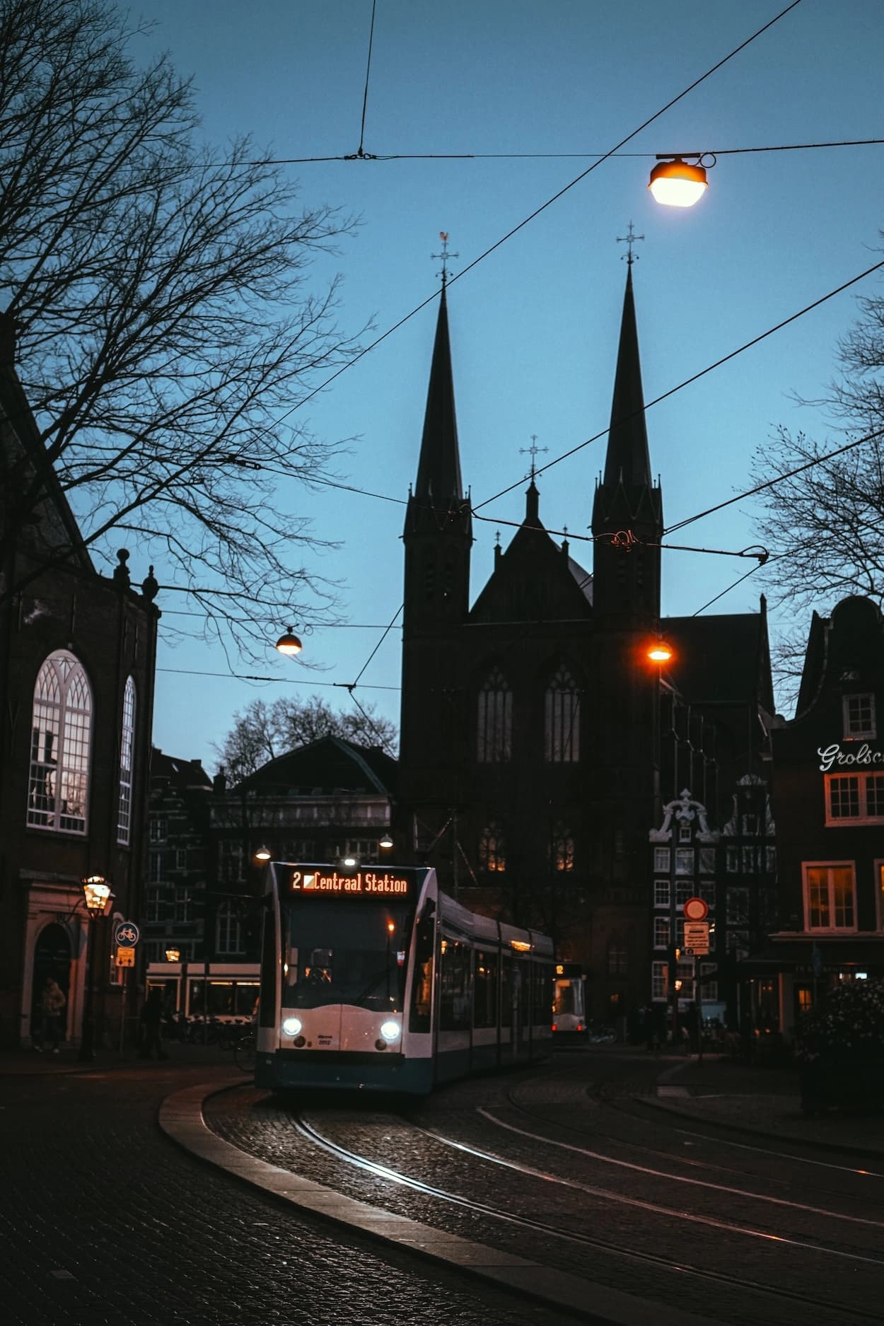 A tram curving over wet cobbles at blue hour, the twin spires of a Gothic church behind it and street lamps burning orange against a deep blue sky