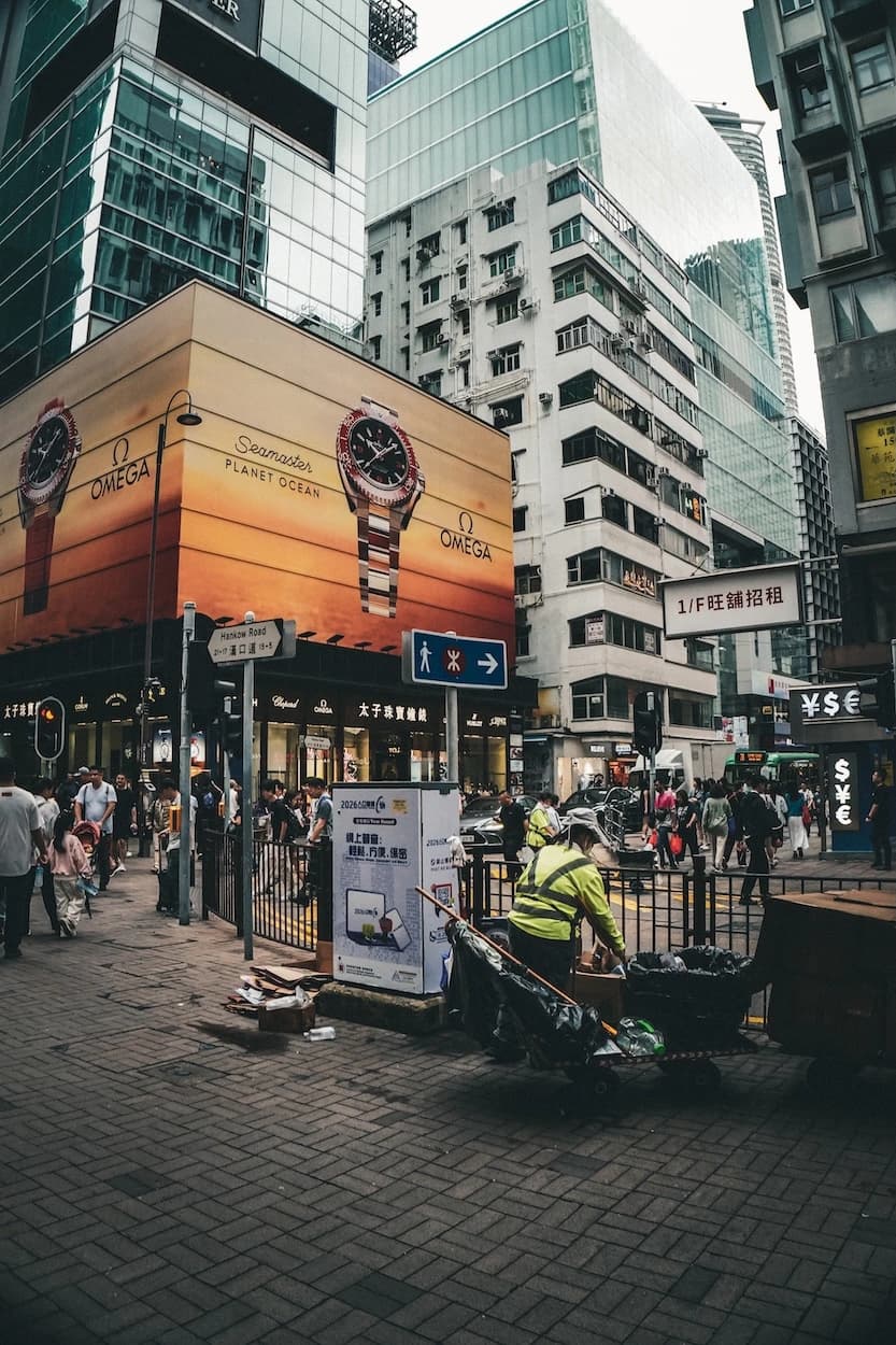 A Tsim Sha Tsui corner at Hankow Road — a giant watch billboard wrapped around a jewellery shop, walk-up blocks and glass towers stacked behind it, and a street cleaner in a yellow high-vis jacket bent over his cart in the foreground