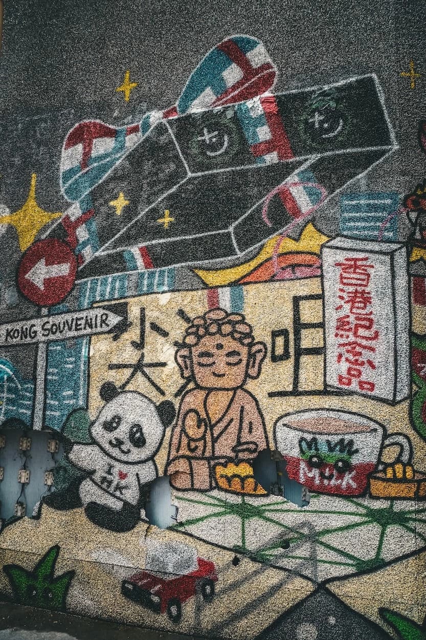 A souvenir-shop mural painted on a rough grey wall — a panda in an I love HK shirt, a seated Buddha, a carton of milk tea and a toppled KONG SOUVENIR street sign against a cartoon skyline