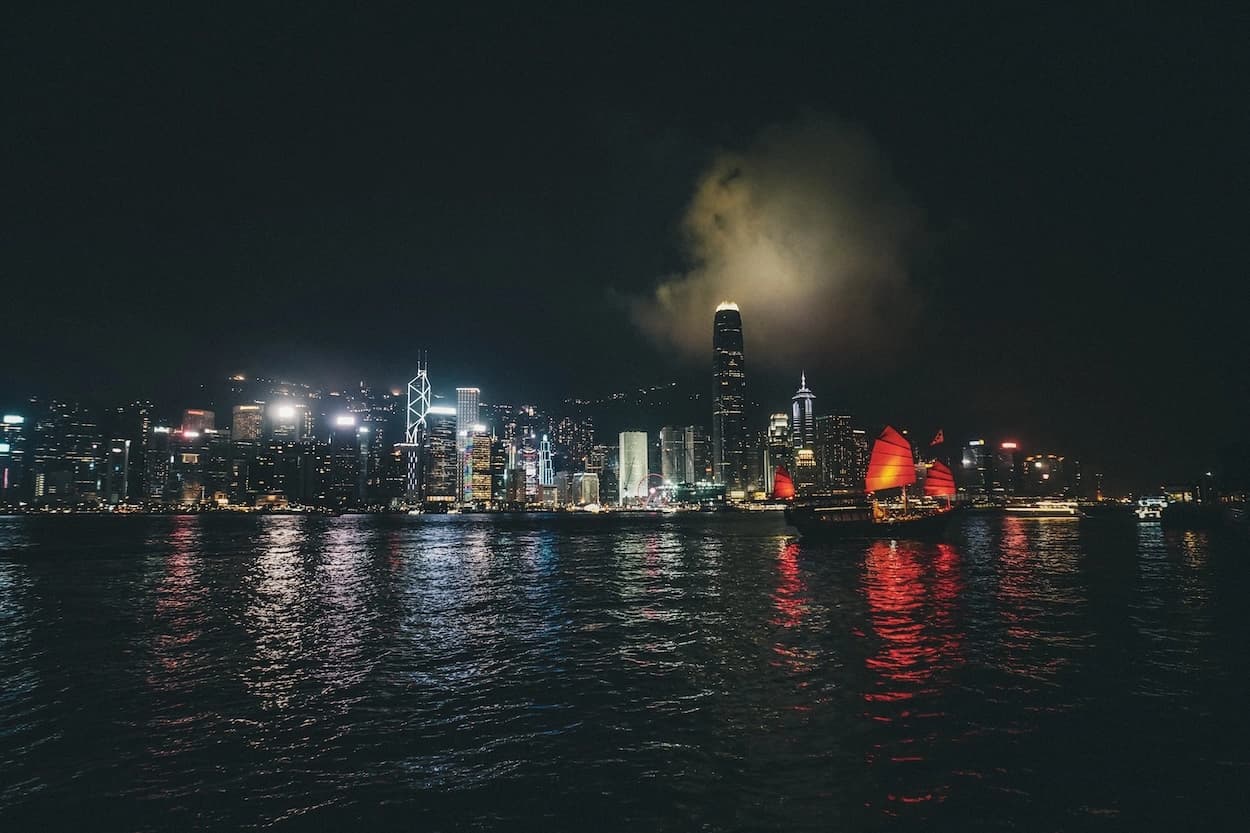 Victoria Harbour at night — a red-sailed junk crossing the water with its reflection running towards the camera, the lit towers of Hong Kong Island stacked along the far shore under a single cloud