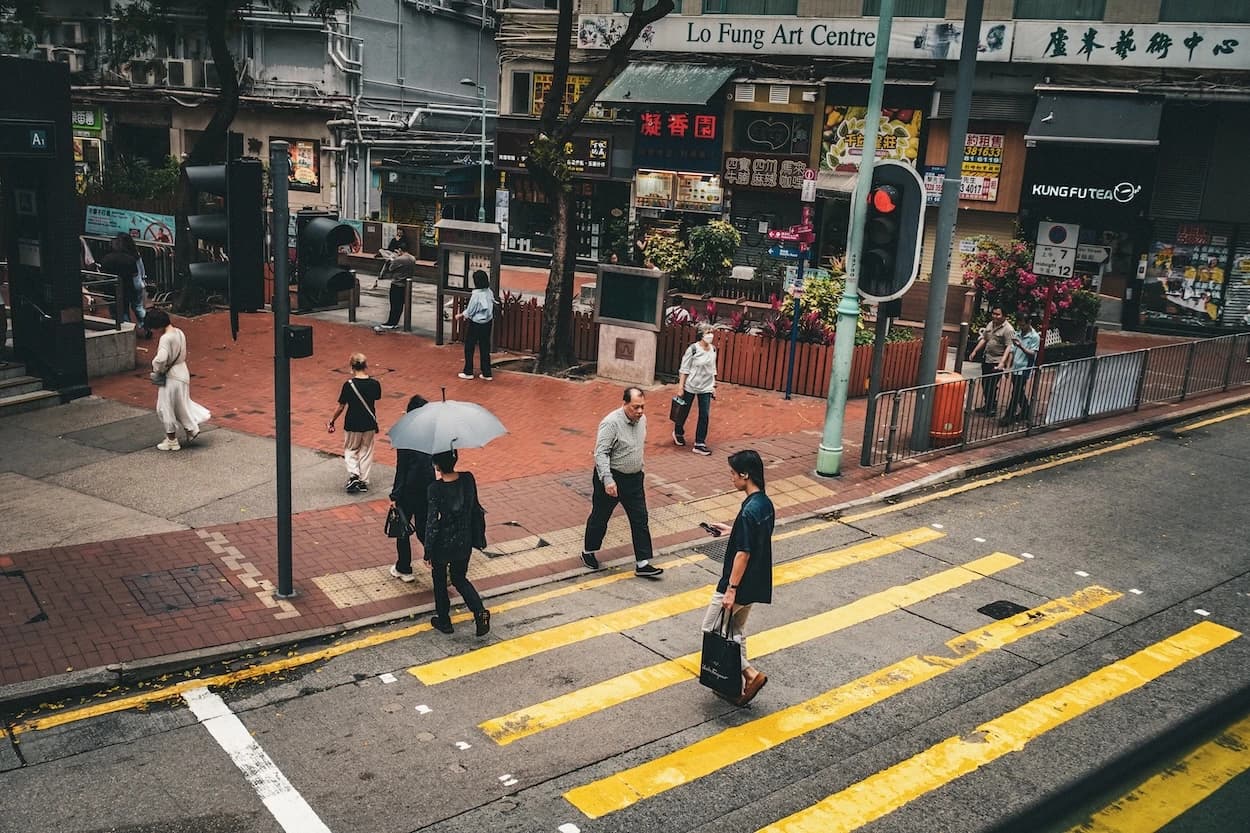 Looking down on a Hong Kong crossing as a man in a black tee carries a paper bag over the yellow stripes, a woman with a pale umbrella behind him and shopfront signs stacked above the pavement