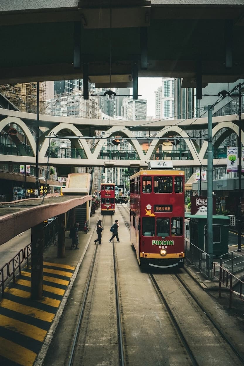 A red double-decker tram waiting on the tracks beneath an arched pedestrian bridge, a second tram further up the line and towers rising behind them into a white sky