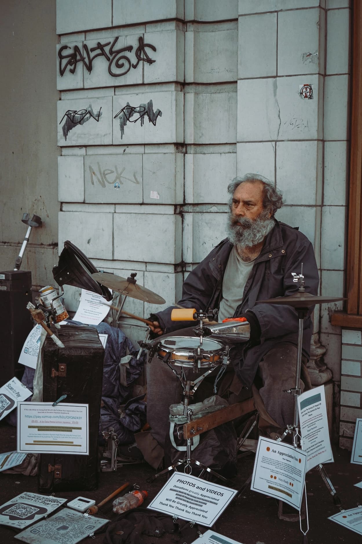 A grey-bearded street drummer mid-beat at a small kit set up against a stone wall, surrounded by handwritten donation signs and cases