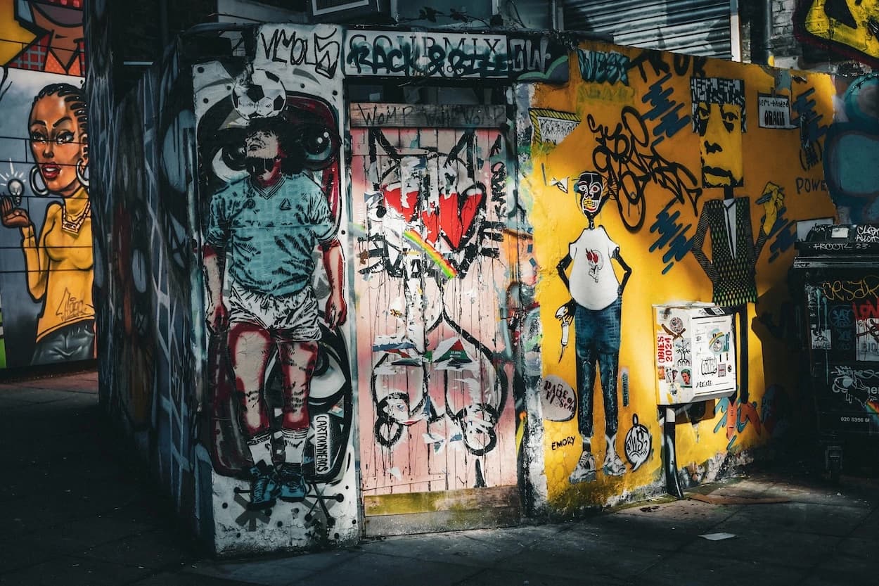 A corner of painted walls and a boarded doorway in an East London alley, a footballer mural on one face and a yellow wall with a stencilled figure on the other