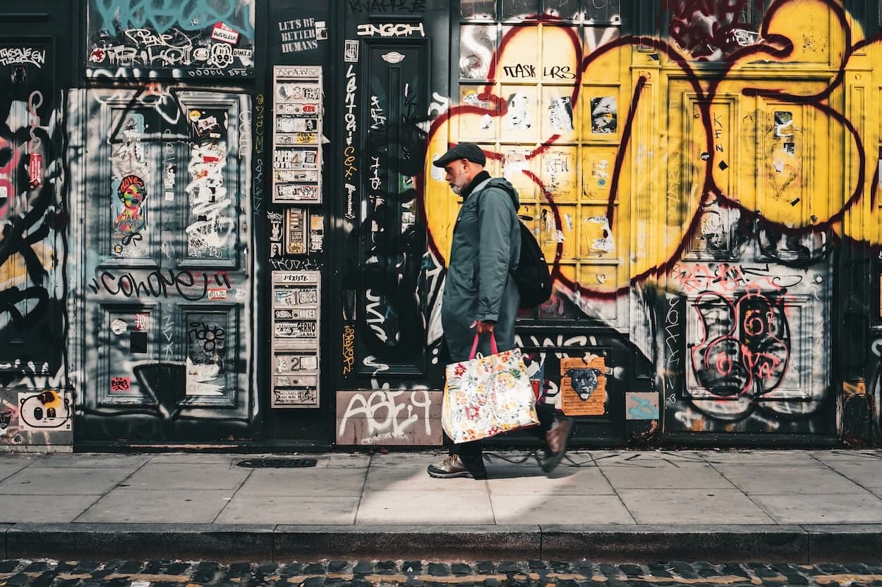 A man in a cap and dark parka walking a Shoreditch pavement past a shuttered shopfront covered in yellow tags, stickers and flyposters, a floral tote bag in his hand