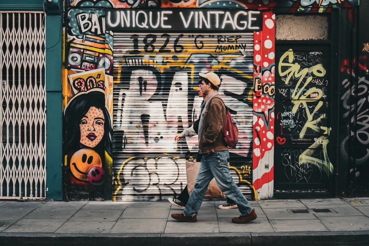 A man in a tan jacket and cap, a paper bag in one hand and a red backpack on his shoulder, striding past a shuttered Shoreditch shopfront painted end to end with graffiti — a pop-art face, a smiley, and a UNIQUE VINTAGE sign above the shutter