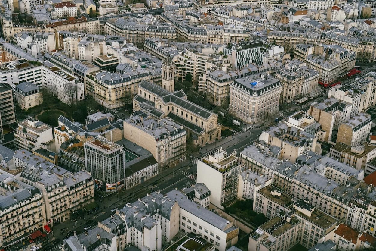 Paris from above — a dense field of zinc rooftops and Haussmann facades running to the horizon, a stone church and its bell tower sitting at the centre of the block