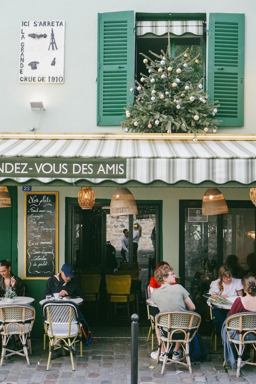 The mint-green front of the Rendez-vous des Amis cafe, a decorated Christmas tree filling an upstairs window above the striped awning and people at rattan chairs on the cobbles below