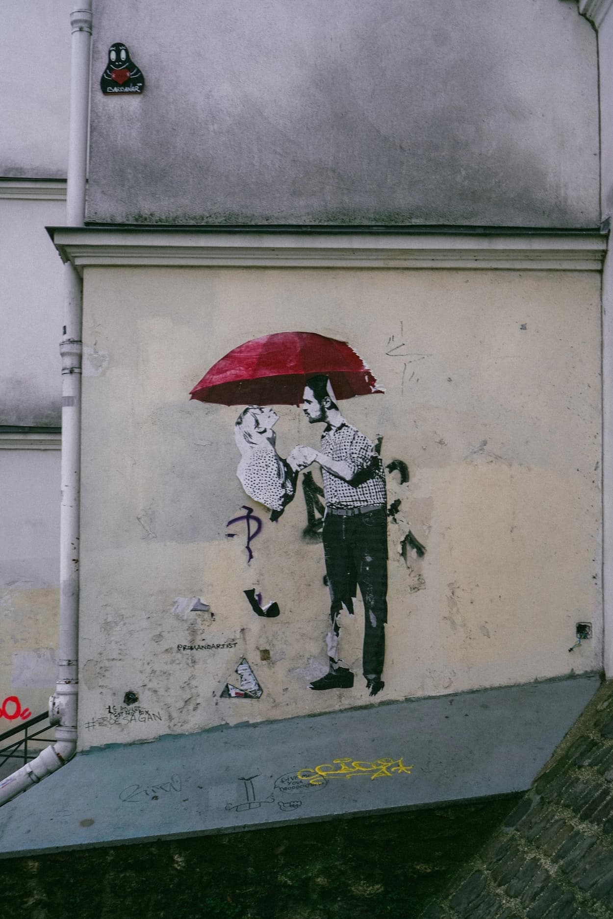 A black-and-white paste-up of a couple under a red umbrella on a weathered Paris wall, a drainpipe running down beside it and tags scrawled across the blue plinth underneath