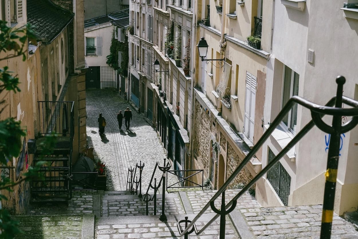 Looking down a Montmartre stairway onto a cobbled lane in low sun, three figures walking through the light between shuttered houses and iron lamp posts